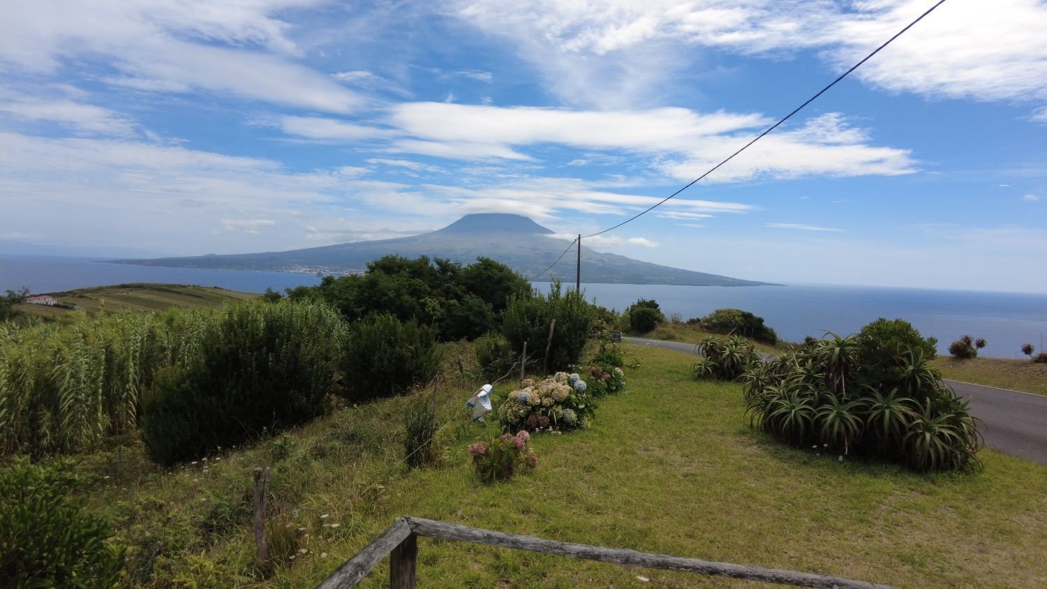 Ein Foto, das von einer leicht erhobenen Aussichtsplattform gemacht wurde. Die untere Hälfte des Bildes zeigt die grüne Landschaft auf der Ostseite der Insel Faial. Im Hintergrund ist zunächst eine Wasserstraße des Atlantiks zu sehen, bevor die Westspitze der Insel Pico zu sehen ist. Zentral von Pico reicht ein Vulkan bis in die Wolken.