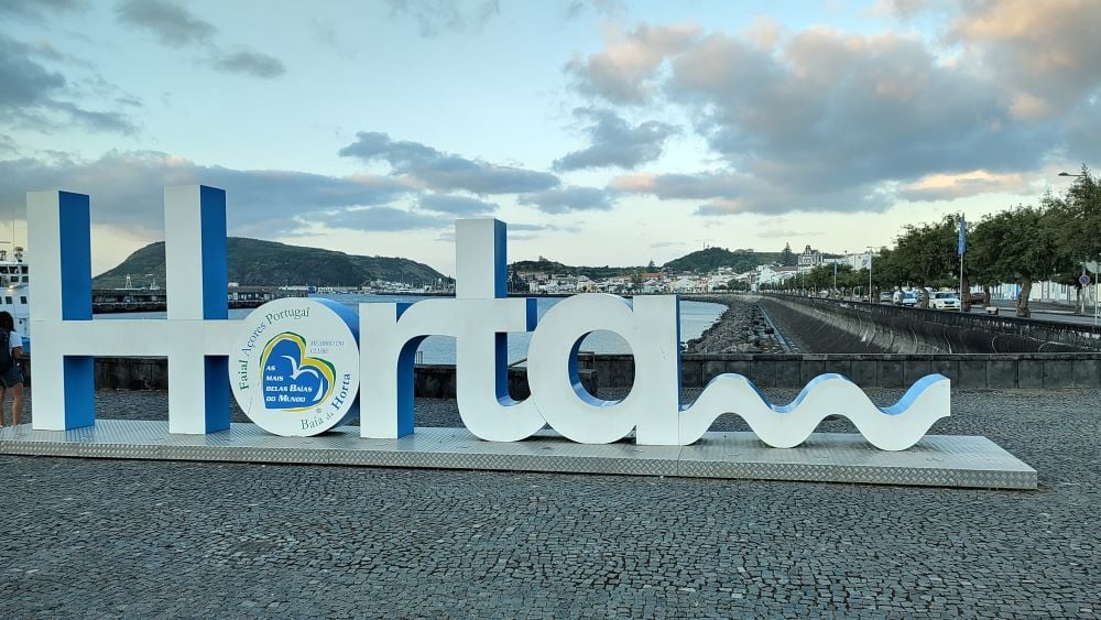 Das Bild zeigt einen Schriftzug aus großen weißen Buchstaben, die vor der Hafenbucht von Horta aufgestellt sind. Der Schriftzug stellt das Wort „Horta“ dar. Im Hintergrund sieht man die Hafenpromenade des Ortes.
