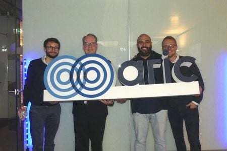 Four men smilingly hold a sign into the camera